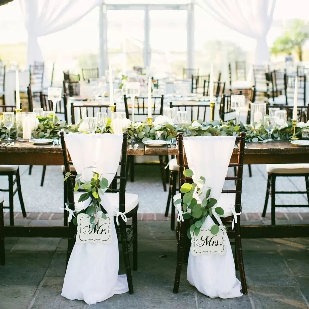 Фото. Деревянные свадебные стулья оформлены с помощью белой ткани и зелени.