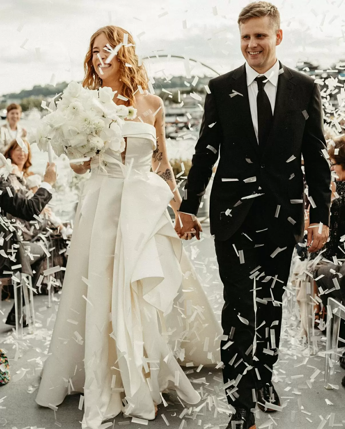Фото Жених и невеста в белоснежном платье с белым букетом в руках идут среди гостей на выездную церемонию и их забрасывают белыми бумажками с пожеланиями.