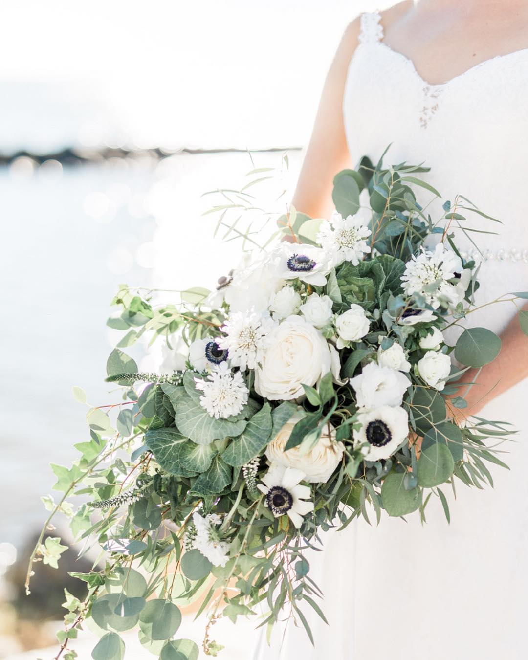 Невеста держит авторский букет с белыми цветами и зелеными листьями.