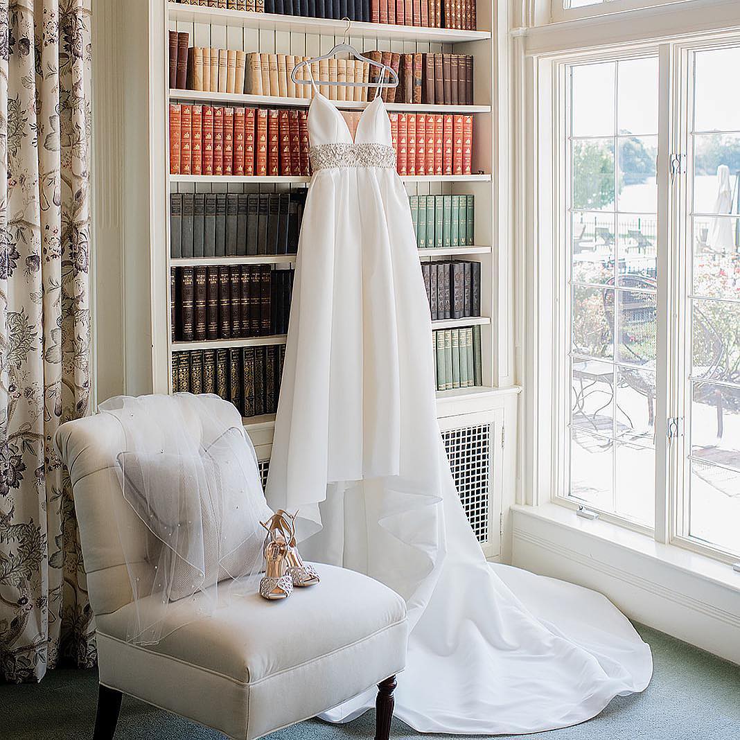 Фото. Свадебное платье висит на вешалке в библиотеке, а на кресле стоят красивые свадебные туфли.
